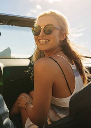Eine junge Frau mit Sonnenbrille auf dem Beifahrersitz eines Cabriolets schaut lachend zu ihrem Partner hinüber, im Hintergrund eine Küstenlandschaft bei Sonnenschein.