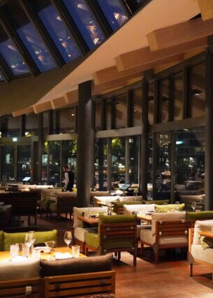 Speiseraum eines modernen Restaurants mit geschwungenem Dach und umlaufender Glasfassade bei Nacht.