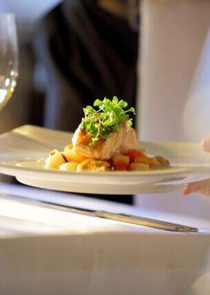 Nahaufnahme eines edel angerichteten Fischgerichtes, das auf einem weißen Teller an einem Tisch mit weißer Tischdecke serviert wird.