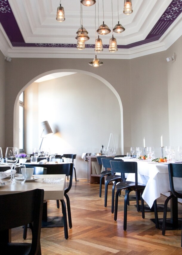 Heller Speisesaal eines edlen Restaurants mit schwarzen Holzstühlen an eingedeckten Tischen in einem klassischen Altbau mit hohen Decken und Holzparkett.