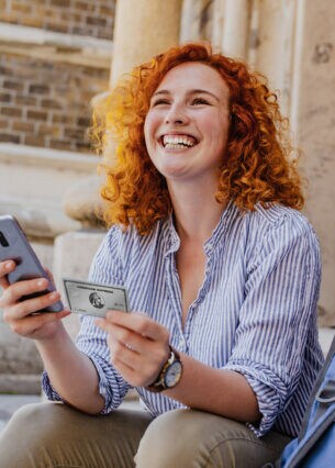 Eine junge Frau mit Smartphone und Kreditkarte in den Händen sitzt gut gelaunt auf einer Treppenstufe am Eingang eines Gebäudes, neben ihr ein Rucksack.
