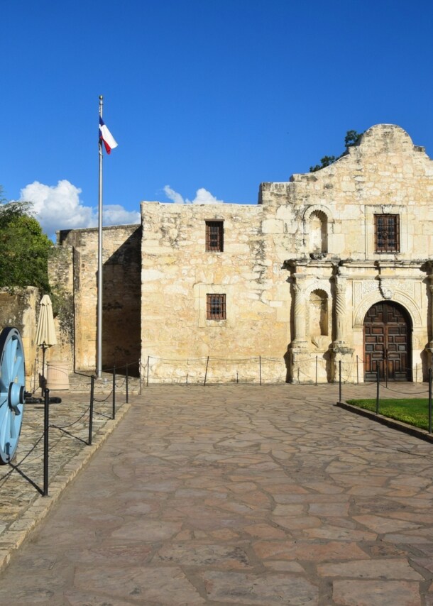 Frontfassade der Kirche einer spanischen Missionsstätte, davor ein Platz mit Texanischer Flagge und einer Kanone.