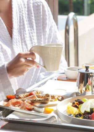 Eine Person sitzt an einem Tisch und hält eine Tasse in der Hand. Vor ihr steht ein Frühstückstablett mit Essen und Getränken.