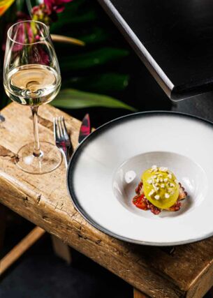 Ein weißer Teller mit einer kreativ angerichteten Vorspeise steht neben einem Glas Weißwein auf einem Holzschemel, daneben ein Blumenbouquet.
