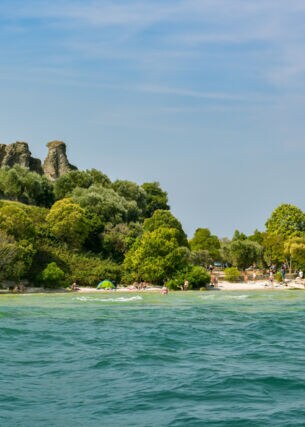 Eine antike Ruine, umgeben von üppiger Natur, an einem Sandstrand mit Personen am türkisblauen Wasser.