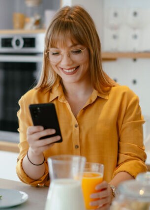 Eine junge Frau in gelbem Hemd steht mit einem Glas Orangensaft in der Hand in der Küche und schaut lächelnd auf ihr Smartphone.