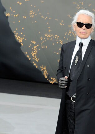Karl Lagerfeld mit Sonnenbrille im schwarzen Jackett winkt vom Laufsteg einer Modenschau.