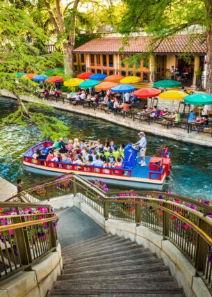 Ein Ausflugsboot fährt auf einem Wasserkanal an einem Restaurant mit Personen auf einer Terrasse mit bunten Sonnenschirmen vorbei, im Vordergrund eine mit Blumen dekorierte Treppe.