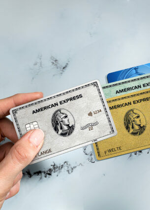Eine Hand hält eine Platinum Card über eine goldene, grüne und blaue Kreditkarte von American Express.