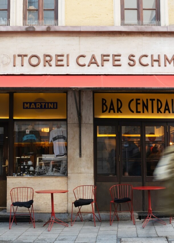 Außenansicht der Bar Centrale mit roter Markise sowie Bistrotischen und Stühlen.