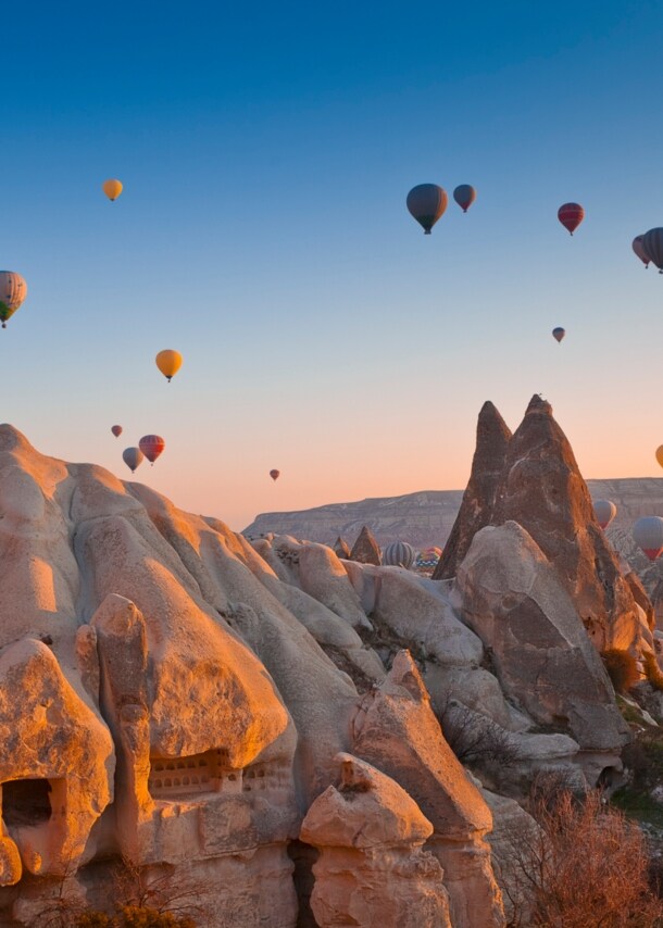 Felsige Landschaft in Kappadokien bei Sonnenuntergang mit zahlreichen schwebenden Heißluftballons im Hintergrund.