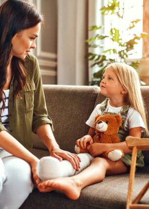 Ein kleines Mädchen mit eingegipstem Bein und Teddybär auf dem Schoß wird von ihrer Mutter auf dem Sofa getröstet