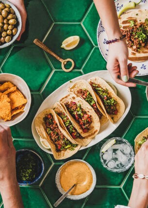 Aufnahme eines Tisches von oben, an dem mehrere Personen sitzen. Auf dem Tisch stehen verschiedene Teller mit mexikanischem Essen wie Tacos, Dips, Nachos und mehr.