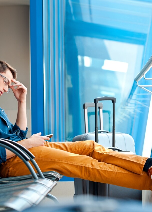 Ein Mann, der am Flughafen auf einer Bankreihe mit ausgestreckten Beinen sitzt. In einer Hand hält er ein Smartphone, die andere Hand ist an seiner Schläfe. Neben ihm steht ein Koffer.