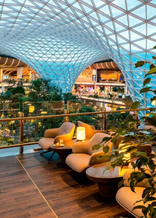 Moderne Lounge auf einer Empore in einem begrünten Flughafenterminal mit Bäumen unter einer geschwungenen Glaskuppel.
