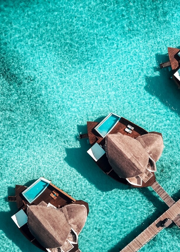 Eine Luftaufnahme einer Hotelanlage mit privaten Suiten samt Pool auf den Malediven, die in einer Linie an einem Steg angelegt und von türkisblauem Wasser umgeben sind
