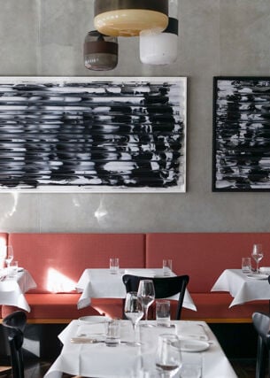Ein lichtdurchfluteter Raum mit mehreren eingedeckten Tischen, weißen Tischdecken, schwarzen Stühlen, einer rot-braunen durchgängigen Sitzbank und zwei großen Bildern mit abstrakten Schwarz-Weiß-Motiven.