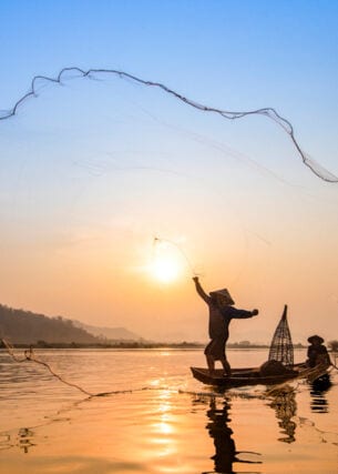 Fischer in einem Holzboot auf dem Fluss Mekong, der bei Sonnenauf- oder Untergang sein Netz auswirft.  