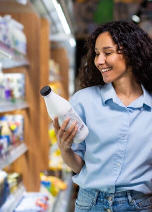 Eine junge Frau steht vor einem Regal in einem Supermarkt und betrachtet lächeld eine weiße Flasche in ihrer Hand.