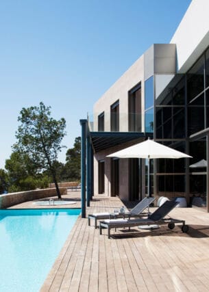 Ein Haus mit großer Glasfensterfront und angeschlossener Holzterrasse mit Pool. Auf der Terrasse befinden sich ein Sonnenschirm und zwei Liegen.