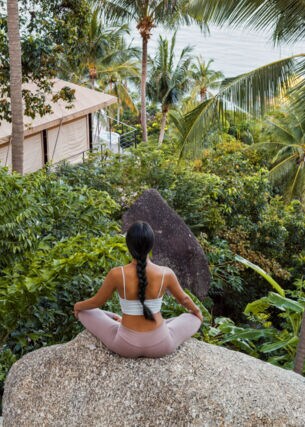 Eine Frau sitzt im Lotussitz auf einem großen Stein, vor ihr eine üppig-grüne Natur, ein Bungalow sowie der Ozean Thailands.