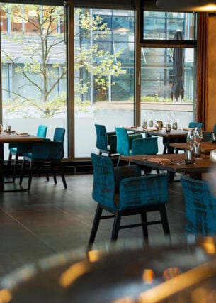 Blaue Polsterstühle an eingedeckten Tischen im Speisesaal eines Restaurants mit bodentiefen Fenstern.