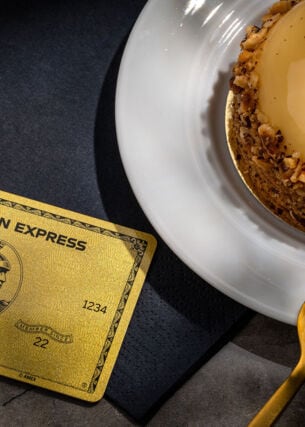 Eine goldene Kreditkarte von American Express liegt auf einer schwarzen Serviette neben einem goldfarbenen Törtchen auf einem Teller und einer goldenen Gabel.