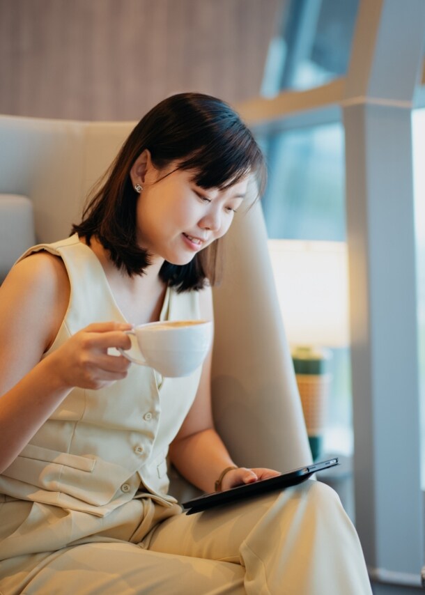 Eine Geschäftsfrau sitzt mit einer Tasse Kaffee in einem Sessel in einer Flughafenlounge und schaut auf ein Tablet in ihrer Hand.