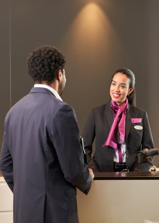 Eine lächelnde Frau in Uniform begrüßt einen Kunden am Empfangstresen einer Flughafenlounge.