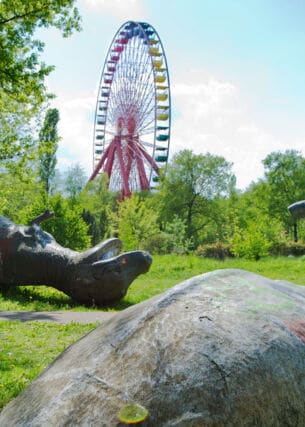 Zwei umgekippte Dinosaurier-Skulpturen auf einer Wiese vor einem Riesenrad in einem Freizeitpark.