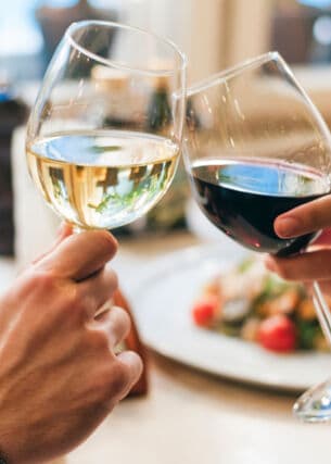 Zwei Personen, die jeweils ein Glas Wein halten und anstoßen. Auf dem Tisch vor ihnen stehen Teller mit Speisen.