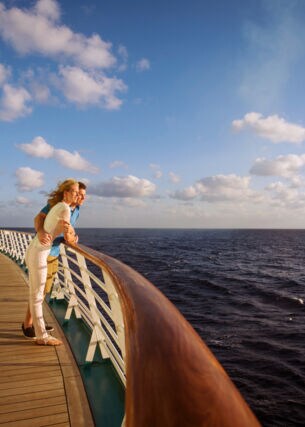 Ein Paar steht an der Reling eines Kreuzfahrtschiffes und schaut aufs Meer im Sonnenschein.
