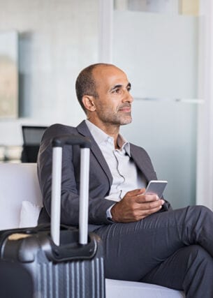 Ein Geschäftsmann sitzt in einem Sessel in einer Flughafenlounge, neben ihm steht ein Koffer.