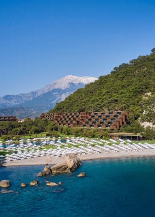 Ein Hotelresort in einer mediterranen Bucht mit Sandstrand, im Hintergrund eine Berglandschaft.
