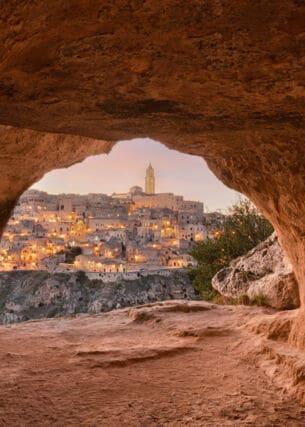 Blick auf die erleuchtete Stadt Matera aus einer Steingrotte im Vordergrund bei Dämmerung.