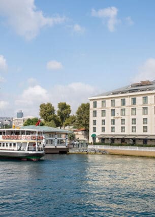 Gebäude mit neoklassizistischer Fassade am Ufer eines Gewässers mit Schiff im Vordergrund, im Hintergrund die Skyline von Istanbul.