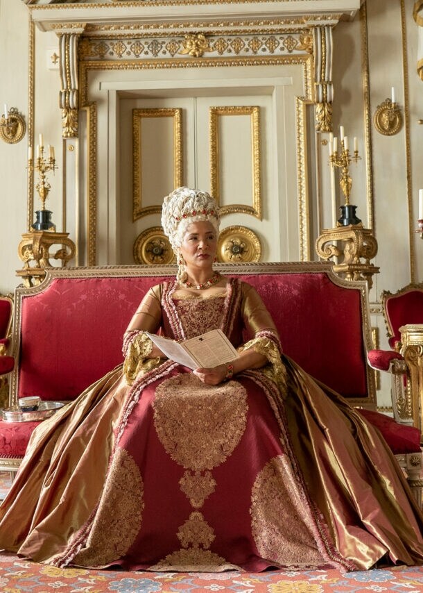 Eine Königin sitzt auf einem roten Samtsofa in einem Palast mit weiß-goldenem Dekor.