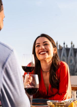 Eine Frau sitzt gegenüber von einem Mann an einem Tisch und hält lächelnd ein Glas Rotwein in der Hnad. Im Hintergrund die Sagrada Familia von Barcelona.
