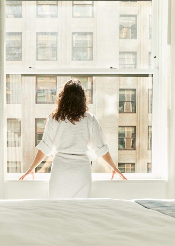 Rückansicht einer Frau im Bademantel, die am Fenster eines Hotelzimmers steht.