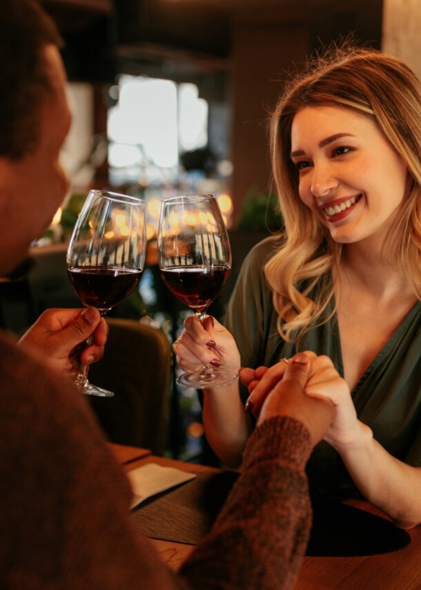 Eine lächelnde Frau sitzt einem Mann an einem Restauranttisch gegenüber und hält seine Hand. In der anderen Hand halten sie je ein Glas Rotwein.