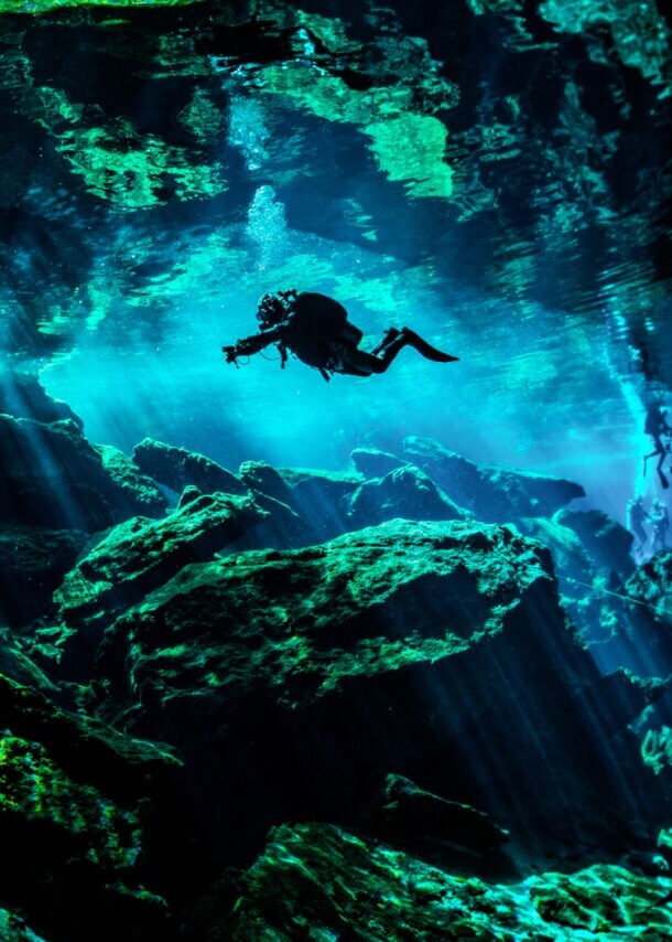 Eine Person in Taucherausrüstung taucht in einer von Wasser gefüllten Höhle. Lichtsäulen ergeben sich durch den starken Lichteinfall von oben im Wasser.