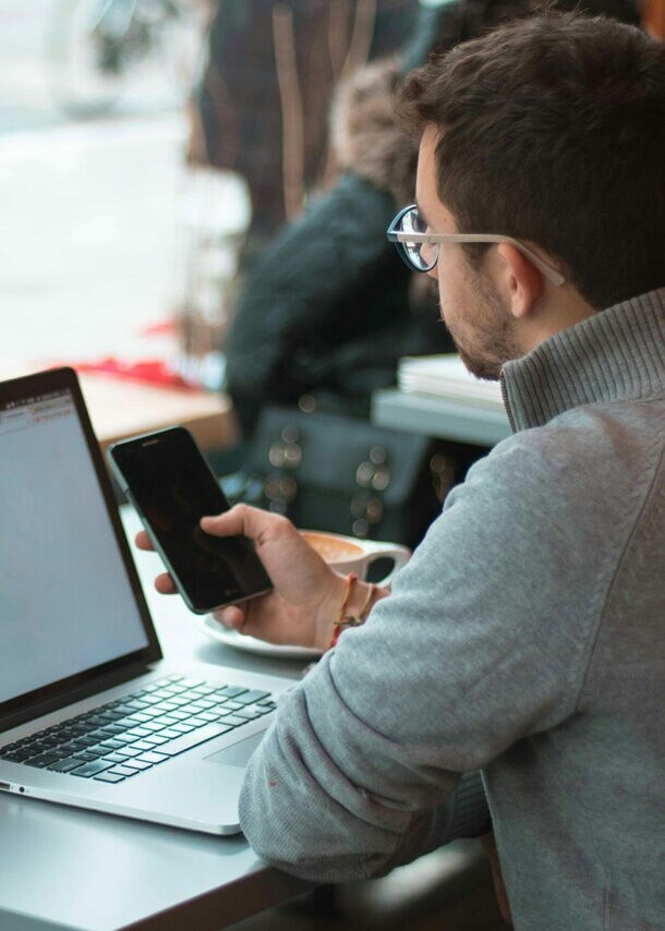 Ein Mann sitzt mit Laptop und im Cafe und schaut auf sein Smartphone.