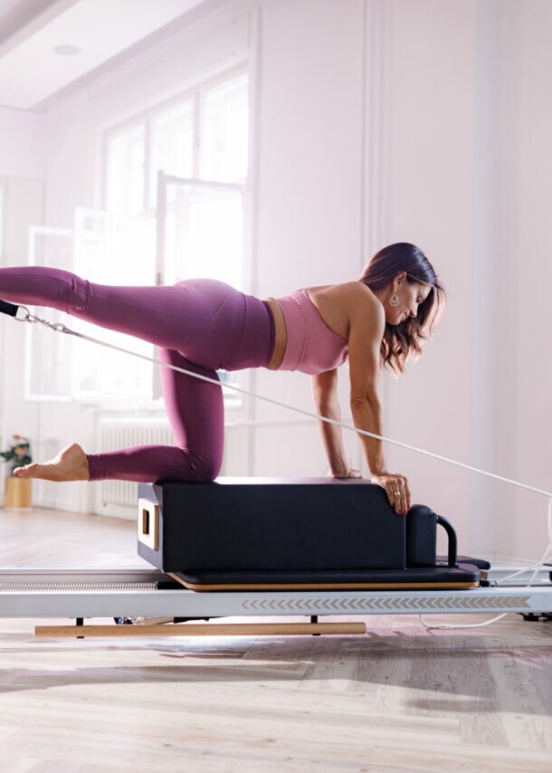 Eine Frau in pinker Sportkleidung kniet auf einer Box auf einem Pilates Reformer in einem hellen Raum.