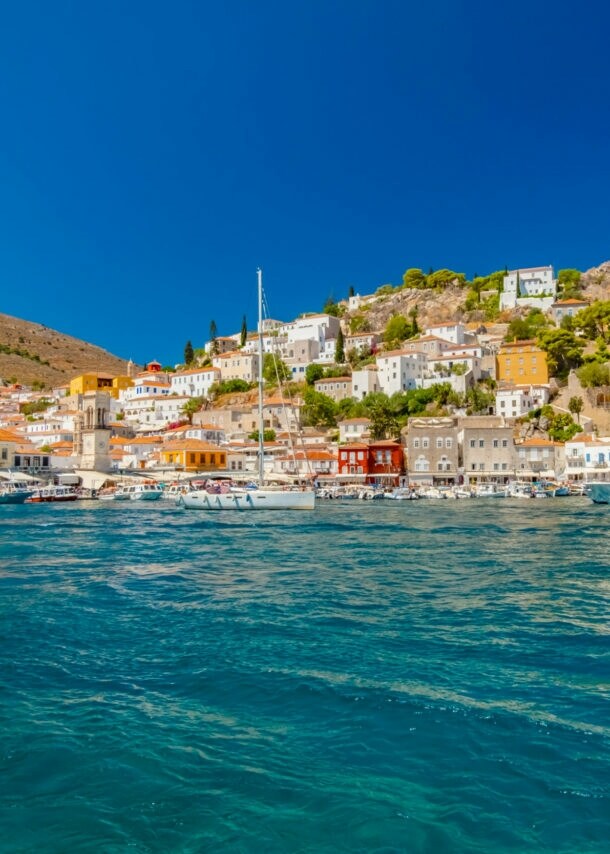 Mediterrane Ortschaft in Hügellage mit Hafenpromenade, im Vordergrund türkisblaues Meer mit Booten.