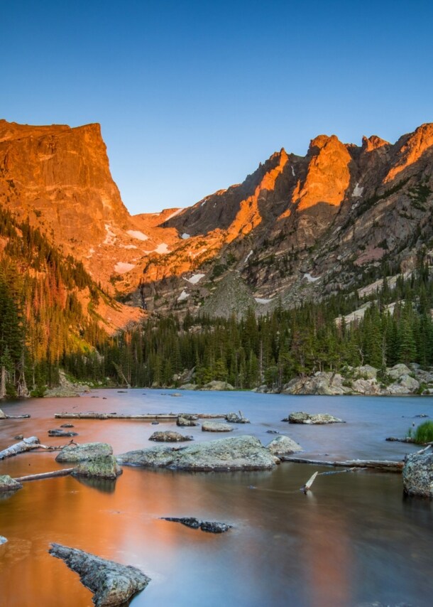 Landschaftspanorama mit See und Nadelwald vor einer schroffen Gebirgslandschaft im warmen Licht bei Sonnenuntergang.