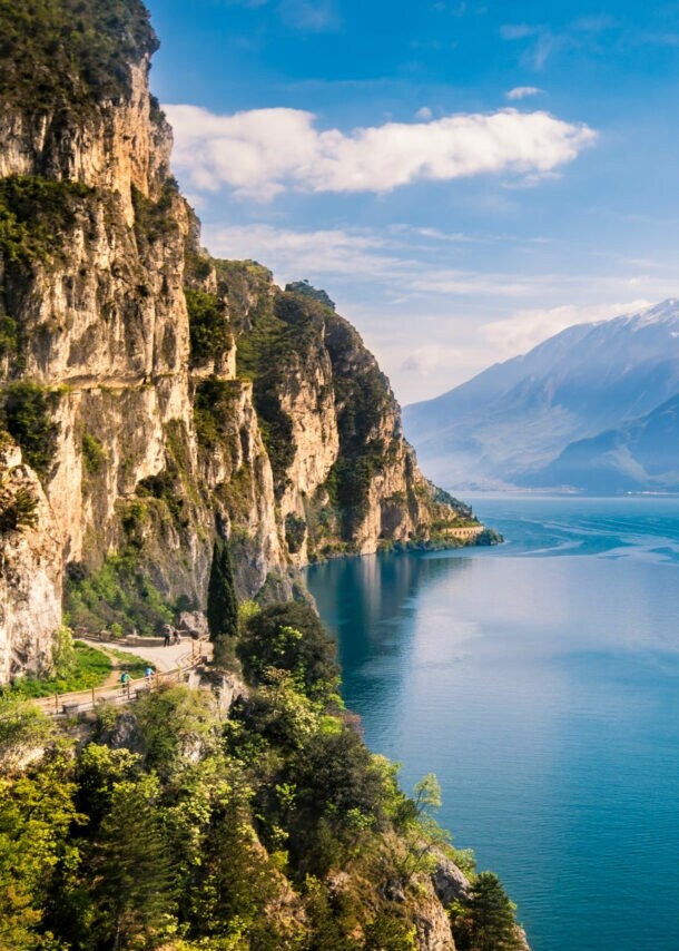 Bewachsene Felswand nahe Riva del Garda direkt am Gardasee mit Gipfeln auf der anderen Seeseite im Hintergrund.