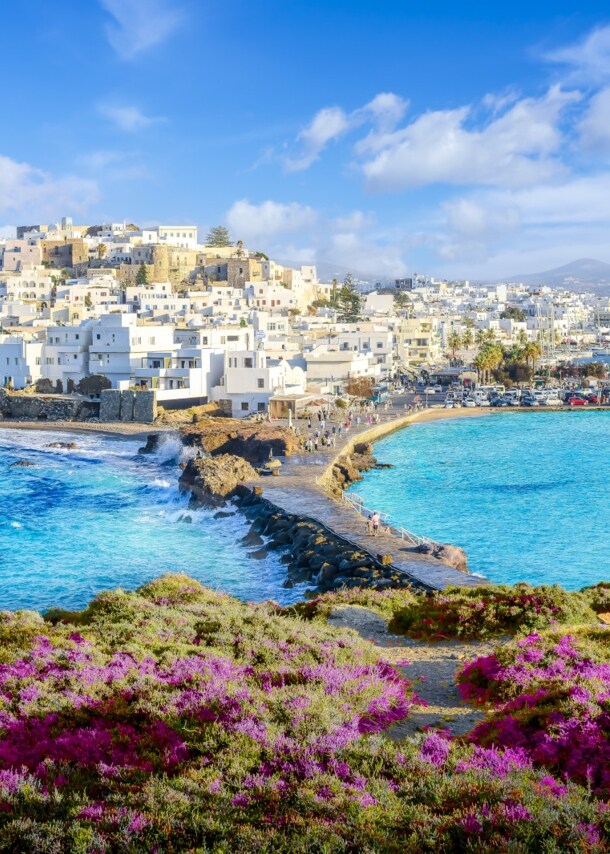 Griechische Kleinstadt mit weißen Häusern und Kaimauer am türkisblauen Meer, im Vordergrund ein bewachsener Hügel.