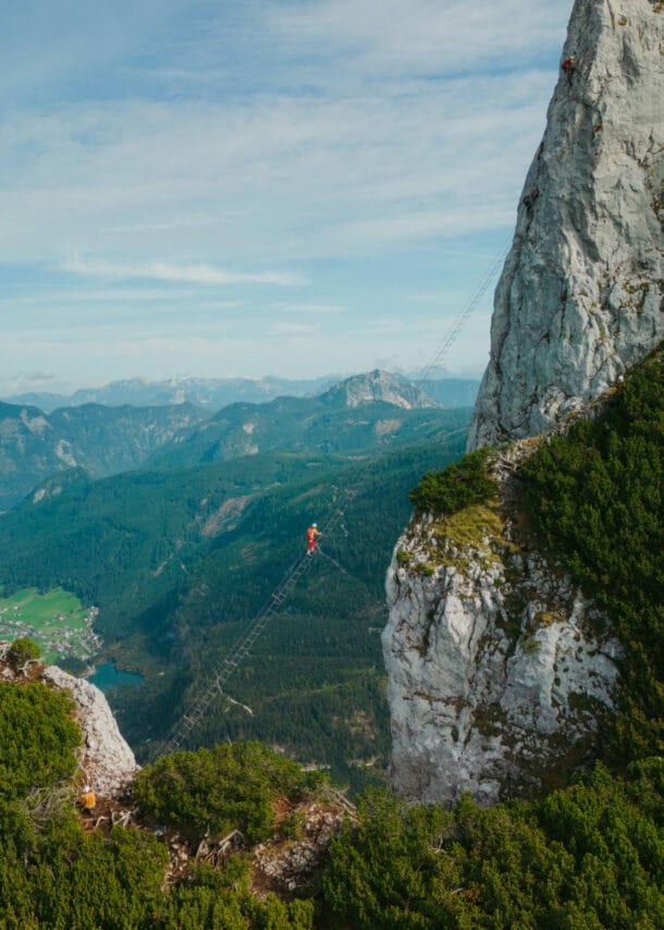 Eine Person auf einer Leiter, die hoch über einem Abgrund zu einem hohen Felsen führt, im Hintergrund Berge und Himmel.