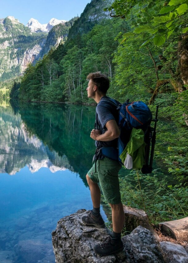 Eine Person mit Wanderrucksack steht auf einem Stein vor dem Königssee mit Blick auf die Berge.