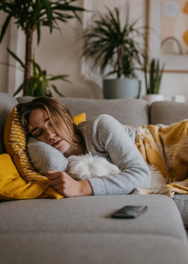 Eine Frau liegt schlafend mit einer Decke auf einer grauen Couch, im Arm hält sie einen Hund.